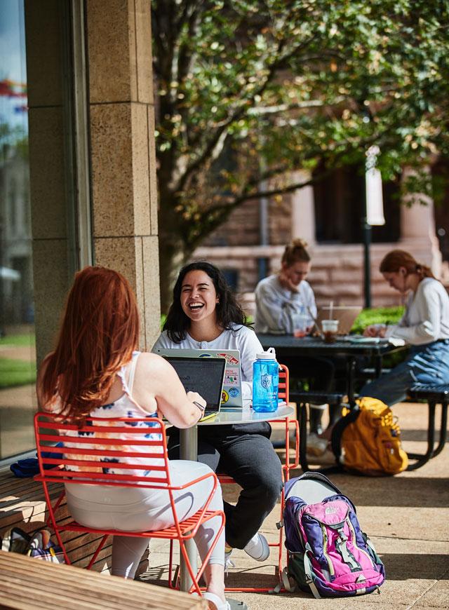 Students at tables outside of the library, smiling and talking while studying